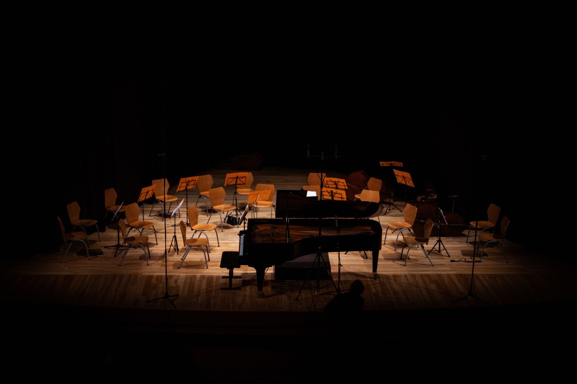 A grand concert hall stage with dramatic lighting — empty chairs and piano await the performers